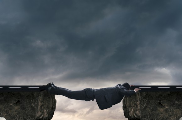 A man in a buisness suit lies across  a gap between two cliffs across a man-sized chasm. 