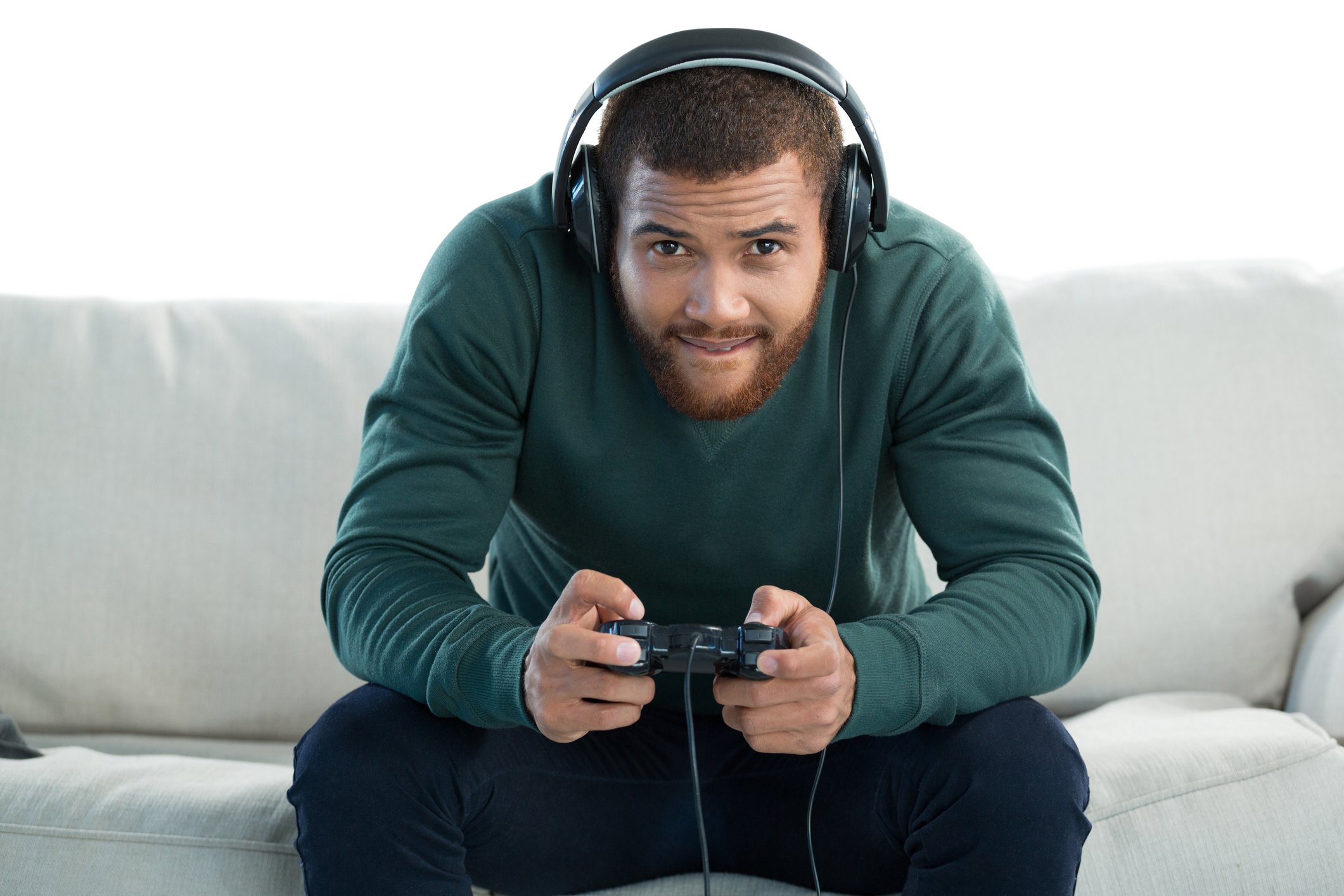 A man wearing headphones and holding a game console controller sits on a white couch, leaning forward to stare at an unseen TV screen.