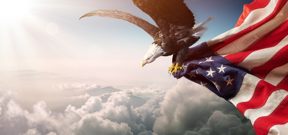 A bald eagle carries an American flag in its claws as it soars through the clouds. 