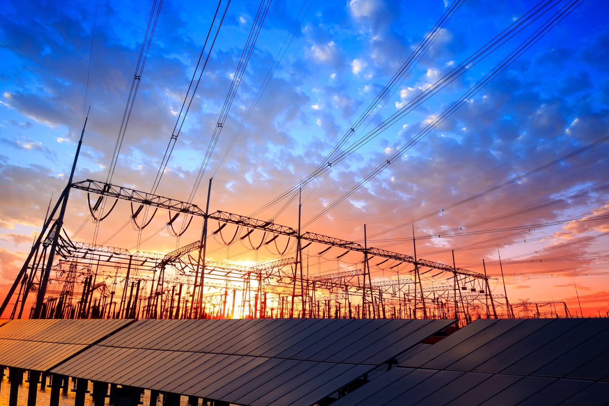 Solar panels and distribution wires stand against the backdrop of a sunset.