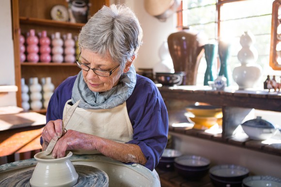 Older person working on a clay pot in a room with other pots and candles.