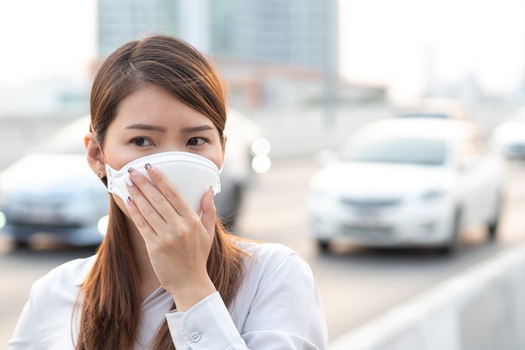 A young Asian woman, wearing a surgical face mask, stands next to a busy city street.