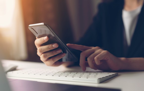 A woman holding her smartphone above a computer