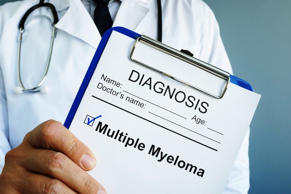 A person in a lab coat with a stethoscope holding a clipboard that has a check mark next to the words multiple myeloma.