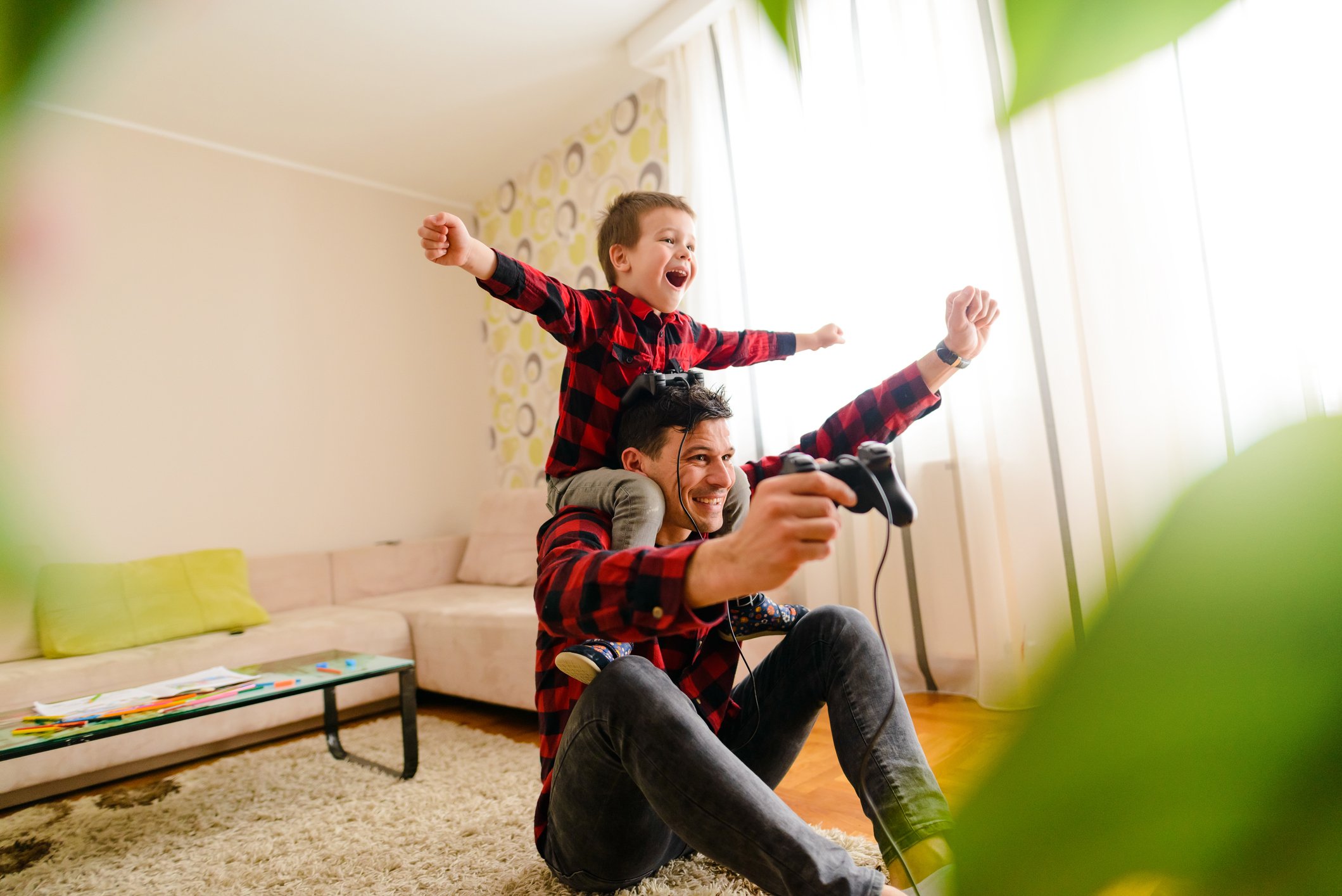 A child on his dad's shoulders cheers his dad, who is playing a console video game in the living room