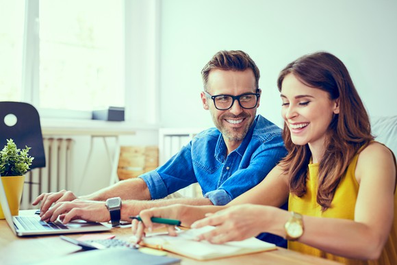 Smiling man typing on laptop next to smiling woman with notepad