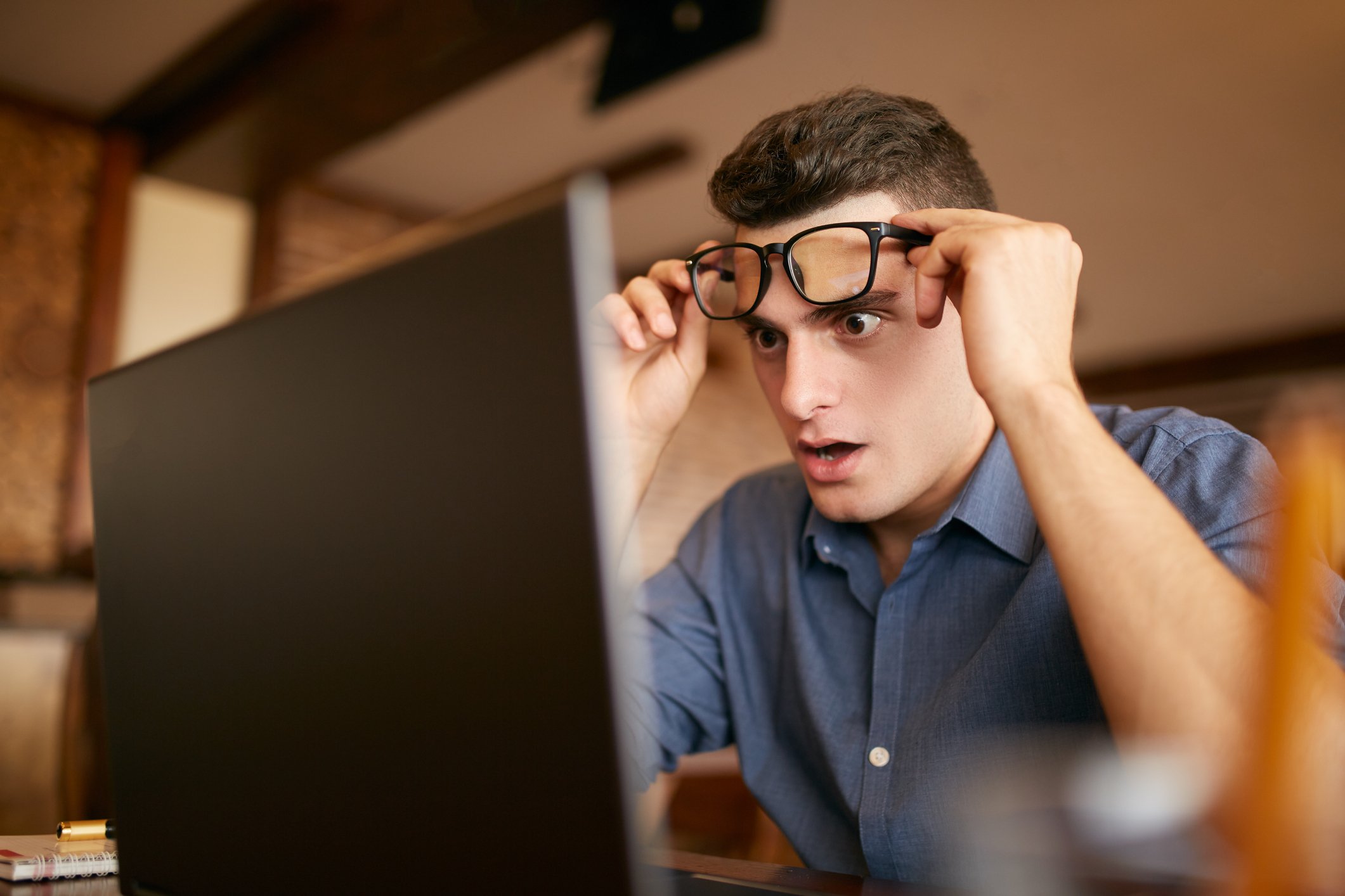 A young man raises up his glasses above his head to look closer at his laptop screen. 