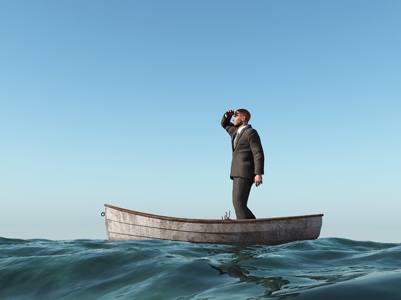 A businessman standing up in a small boat at sea peering out into the distance.