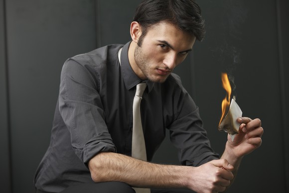 A young businessman glares at the camera, holding a lighter in one hand and a burning piece of paper in the other.