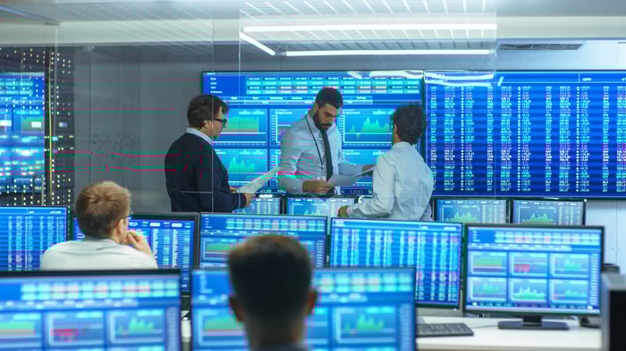 Three people standing in front of room with a dozen big screens showing stock charts and quotes.