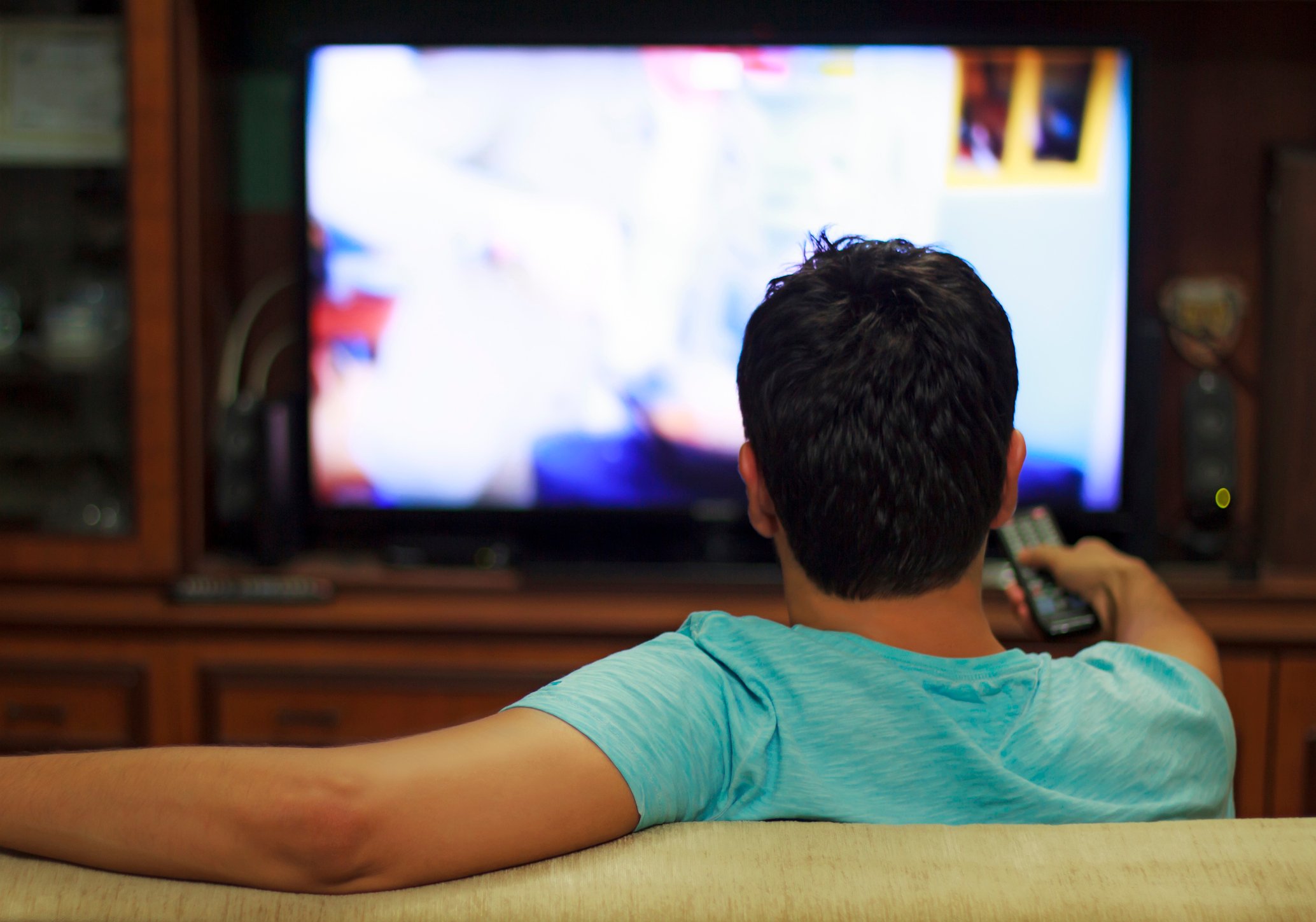 A man sitting on a couch watching TV as seen from behind.