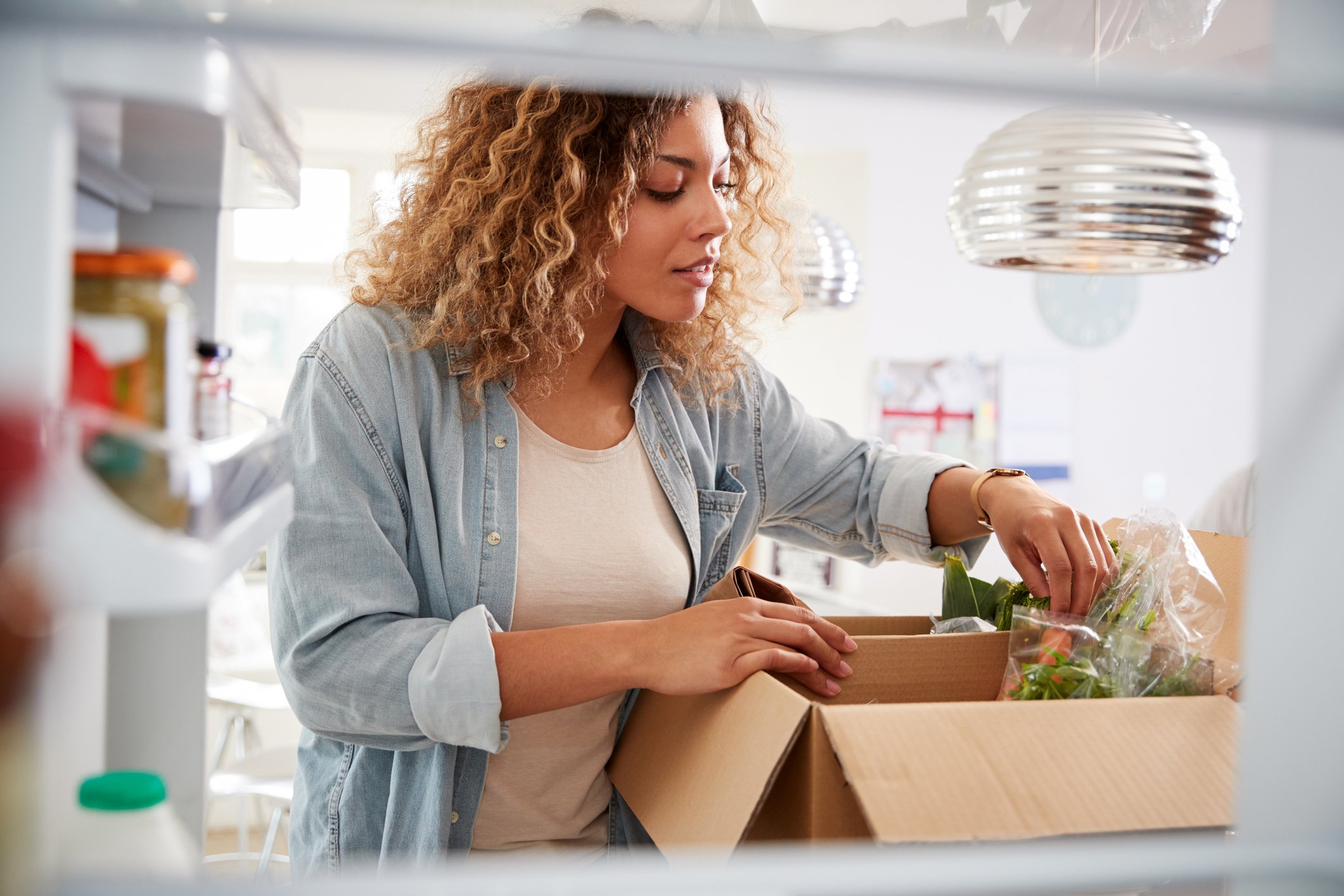 A woman looks into a box containing her grocery delivery as she stands in her kitchen.