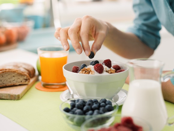 A woman's hand adds a blueberry to a bowl of healthy cereal on a table with milk, juice and a bowl of blueberries.