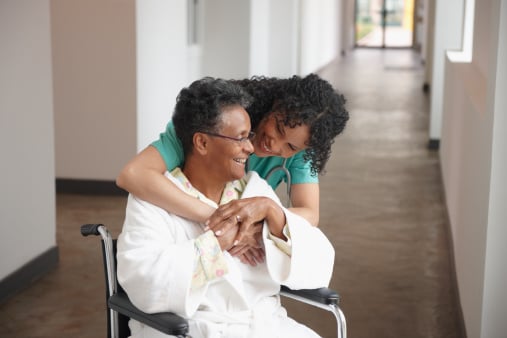 Woman patient in wheelchair being hugged from behind by female healthcare provider