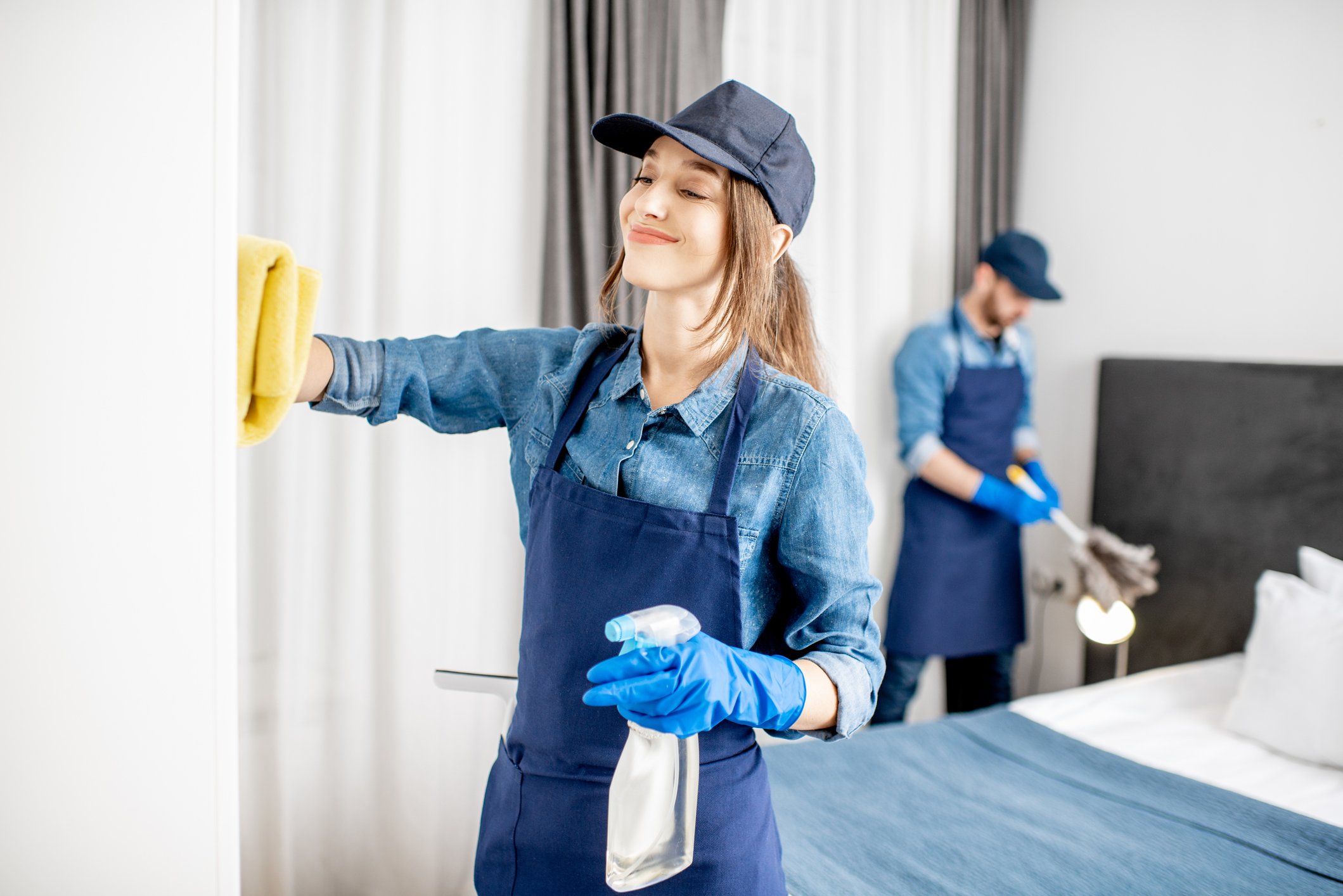 A young woman in uniform cleans a wall with a rag  and a spray bottle of cleanser in the other. 