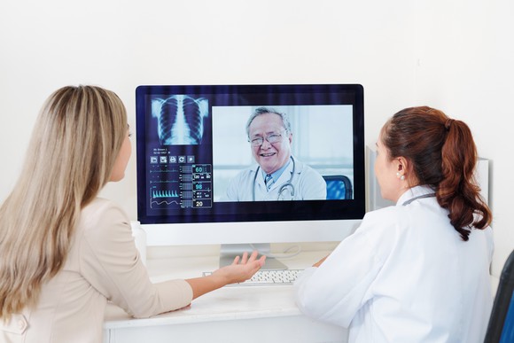 Two young women gather round a computer and speak with a doctor through a video conference via desktop. 