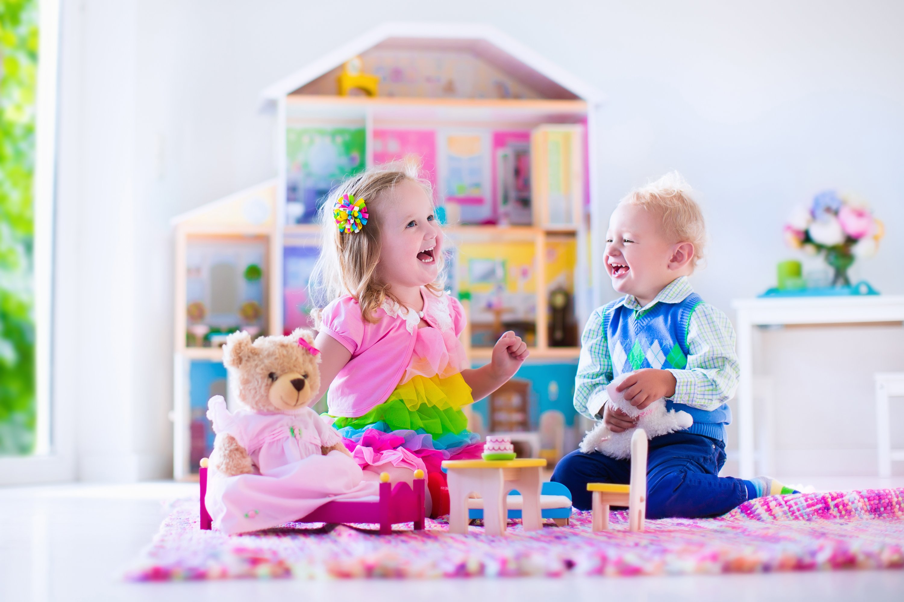 A little girl and boy play teatime with a stuffed bear in front of a doll house.