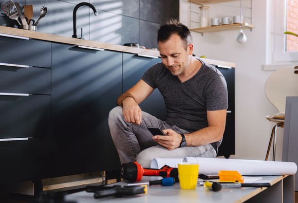 A man kneeling next to his kitchen sink with tools and parts nearby=