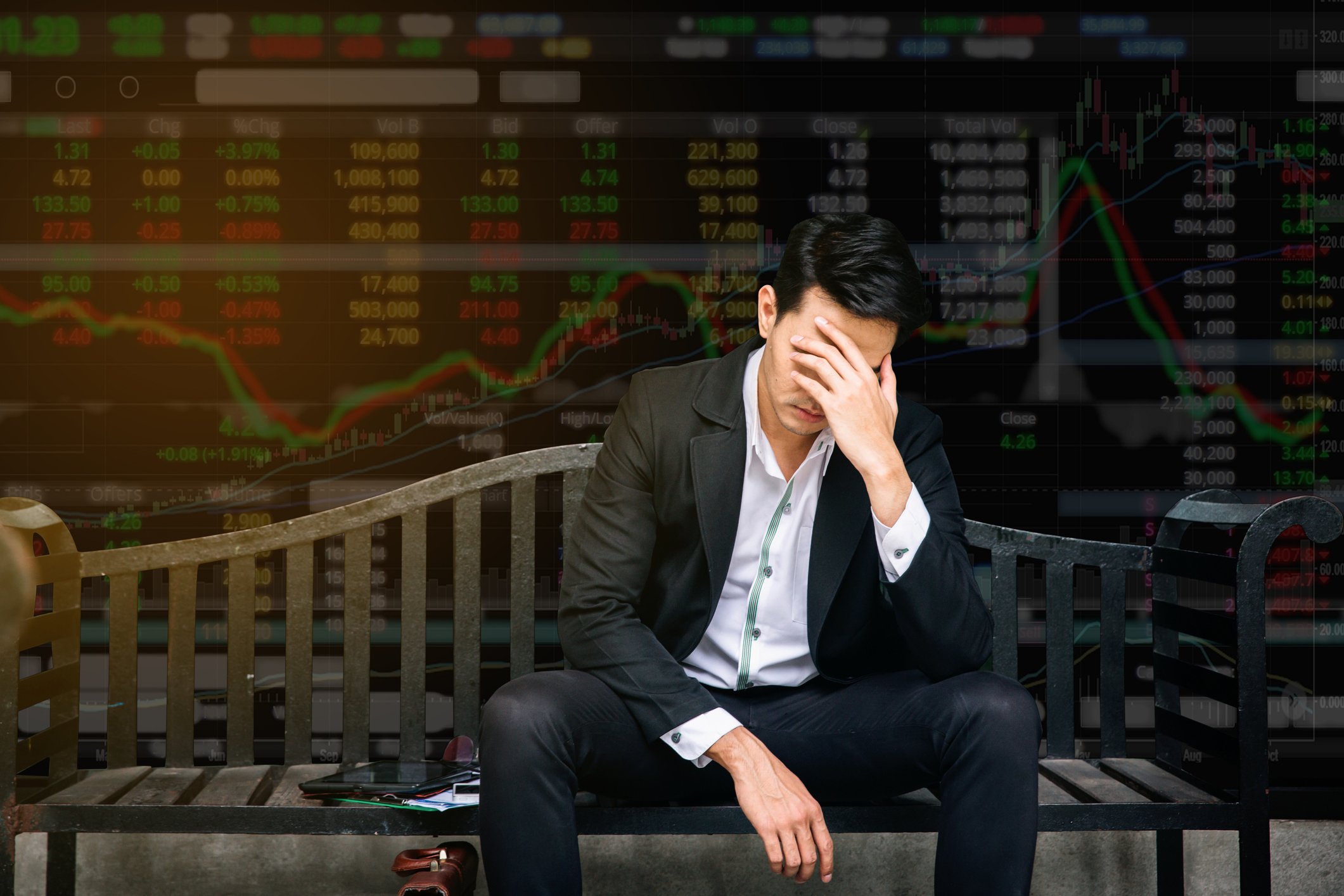 Man in suit sitting on bench with head in hand, in front of a wall displaying numbers and ticker symbols.
