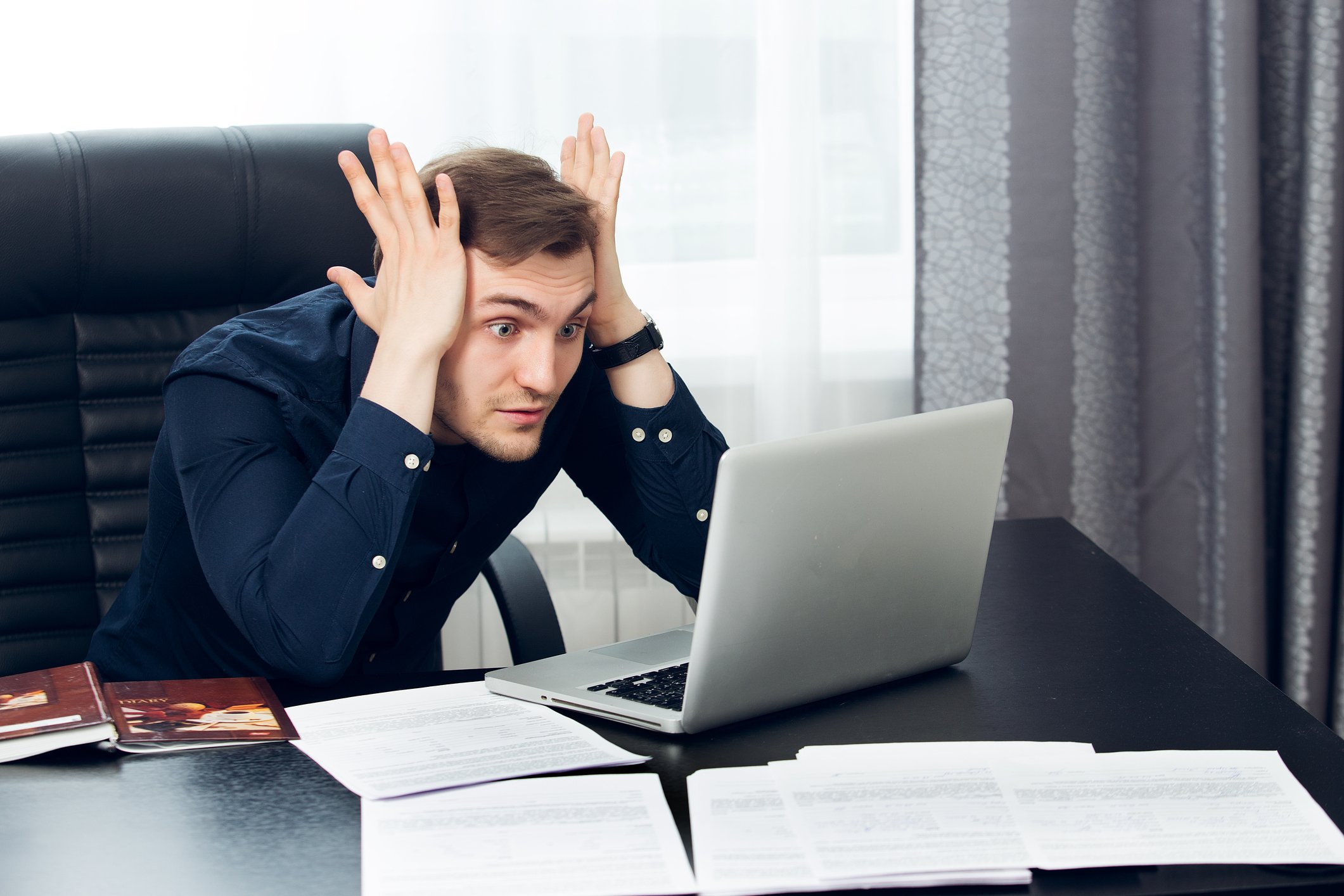 A young man at his desk, staring at his laptop screen with both palms pressed against his temples.