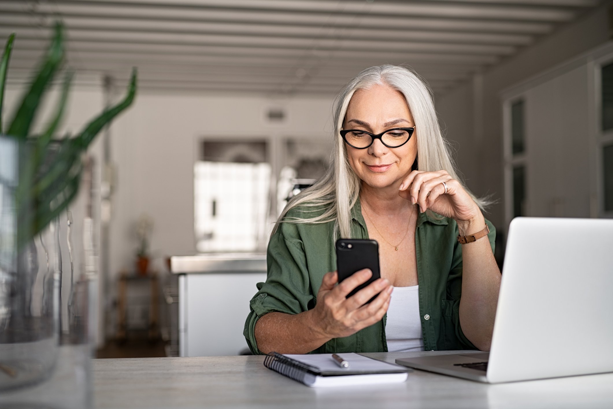 A smiling woman seated at a desk looks at her smartphone.