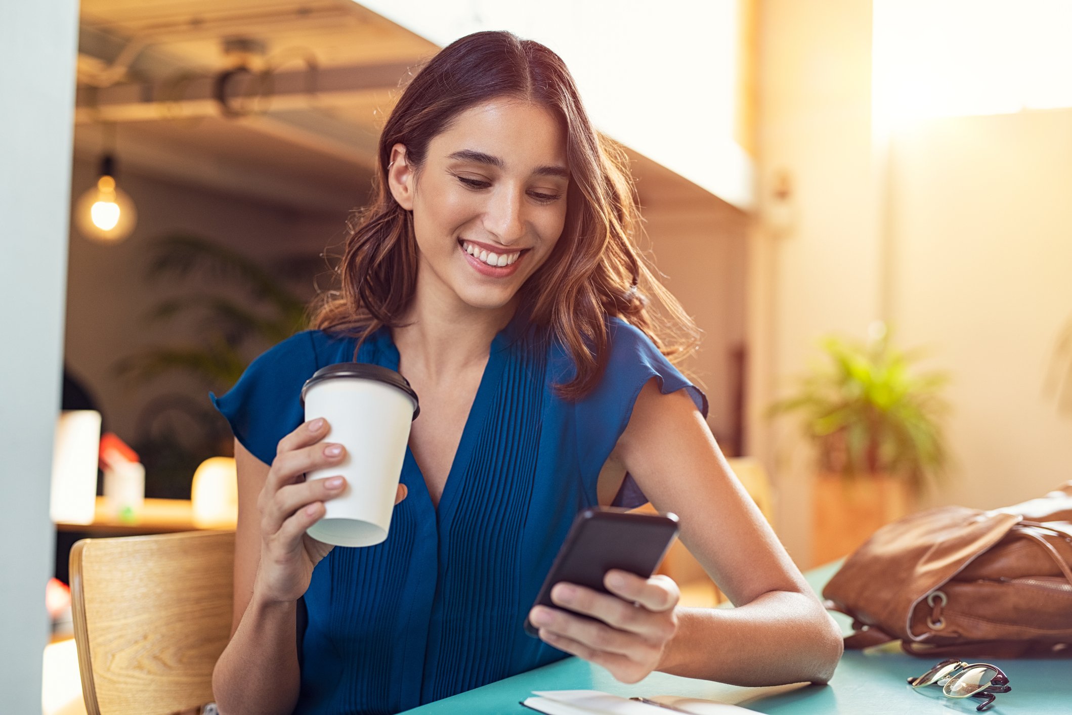 A smiling woman looks at her smartphone while holding a to-go coffee cup.