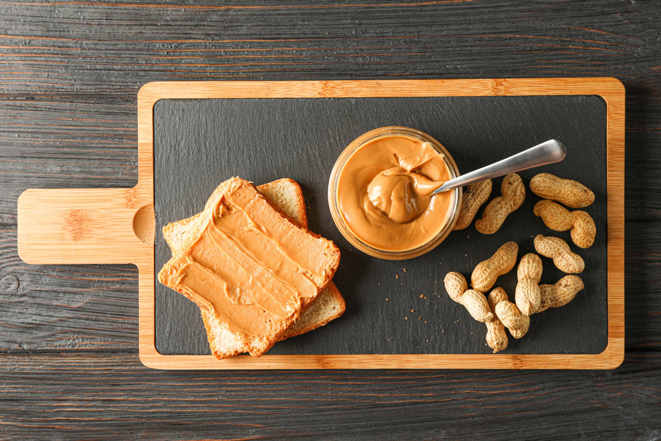 A slice of bread spread with peanut butter is pictured next to a bowl of peanut butter and peanuts on a serving tray.