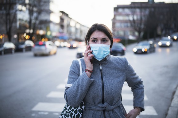 Woman walking across a street, talking on the phone and wearing a face mask.