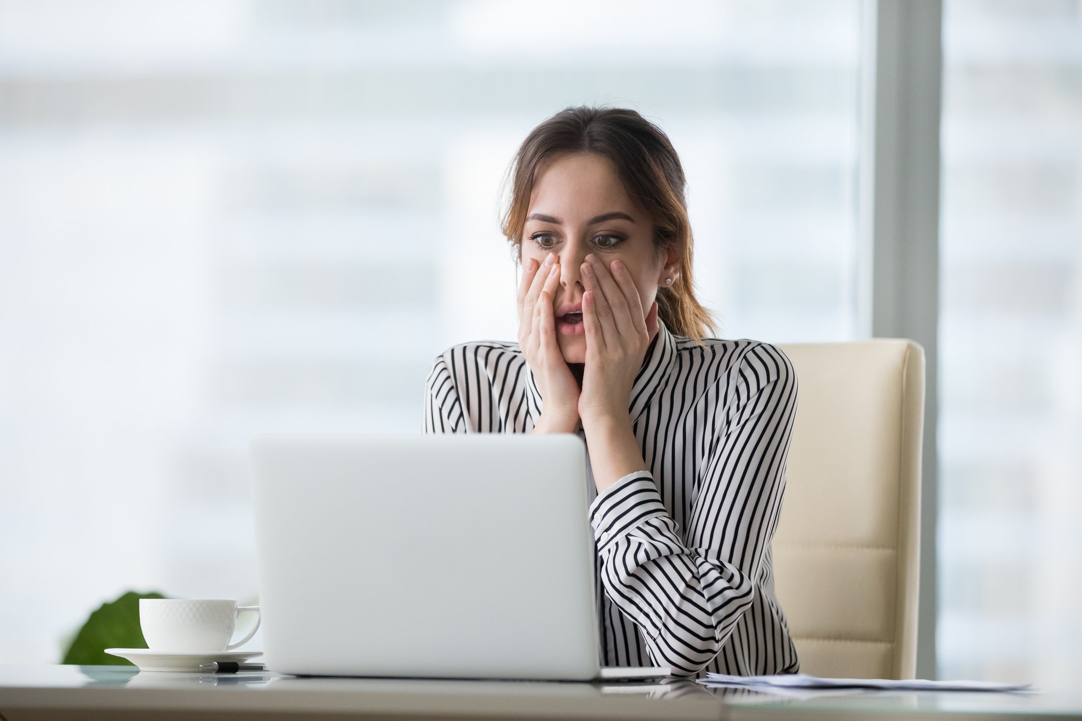 A professional-looking woman staring at a laptop with a look of shock on her face.