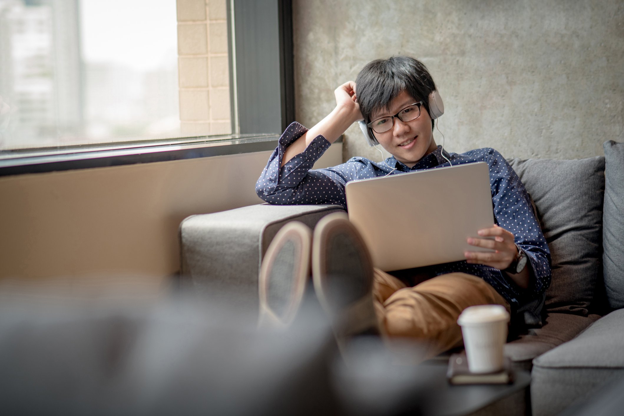 A young asian man sits alone on a couch, smiling at his tablet computer and wearing headphones.
