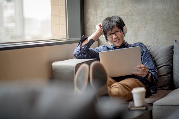 A young asian man sits alone on a couch, smiling at his tablet computer and wearing headphones.