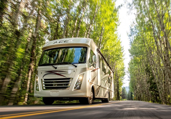A Thor Class A motorhome drives on a road through a clearing of trees.. 