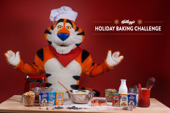Wearing a chef's hat, Tony the Tiger sits behind a table with a variety of Kellogg products