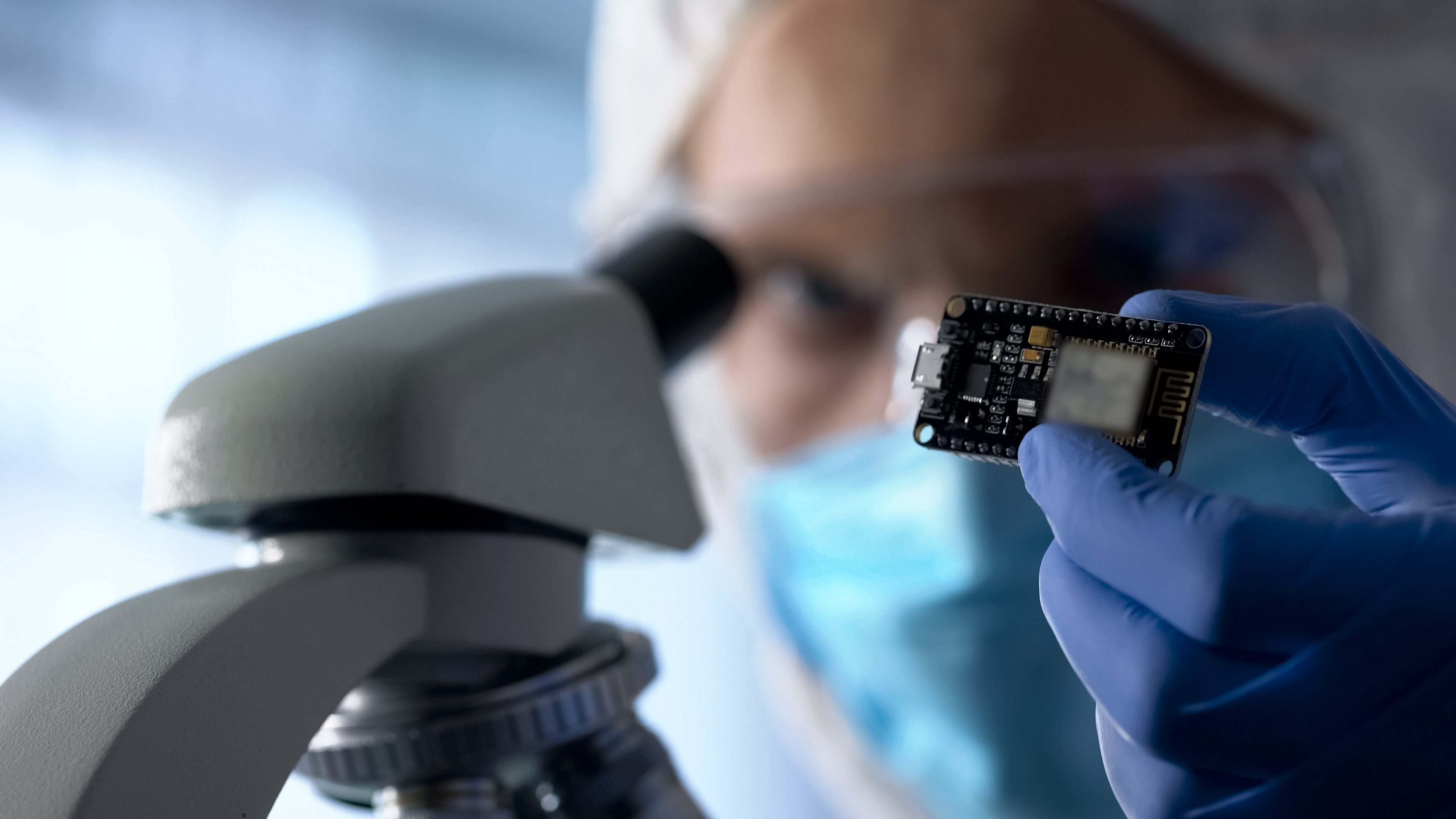 Man in protective glasses holding a semiconductor chip in a blue, gloved hand by a microscope.