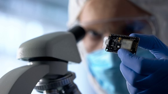 Man in protective glasses holding a semiconductor chip in a blue, gloved hand by a microscope.