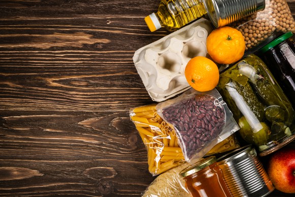 Packaged foods including pasta, oil, and canned goods are shown on a wooden surface.
