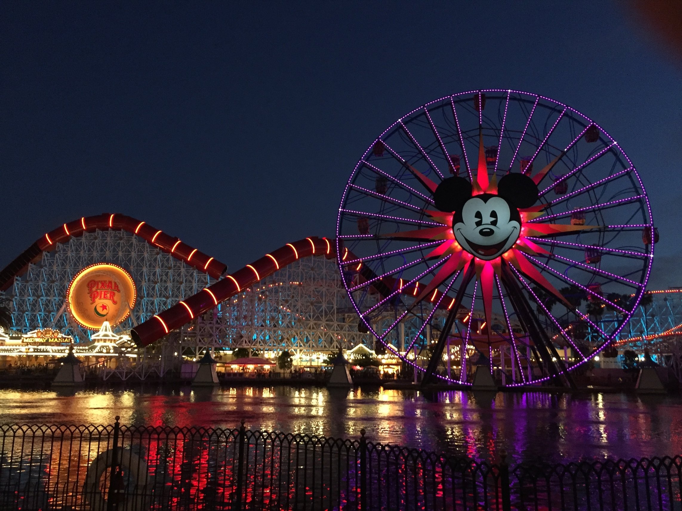 A roller coaster and a Ferris wheel with Mickey Mouse lit up at light.