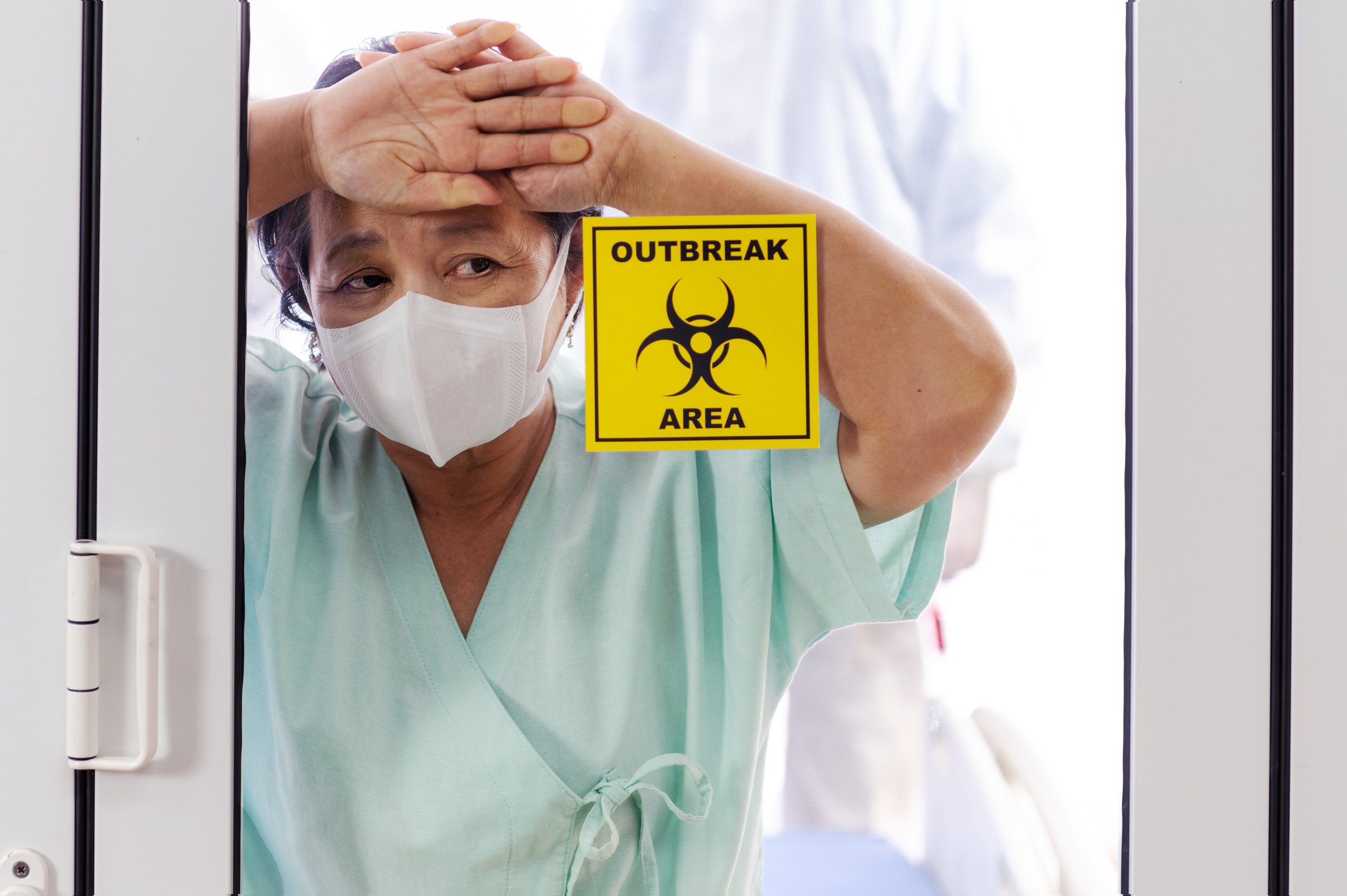 Patient wearing a mask with her hands against a glass door with a sign stating "outbreak area"