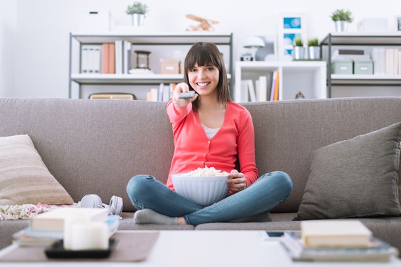 A young woman smiles on the couch and clicks a TV remote.