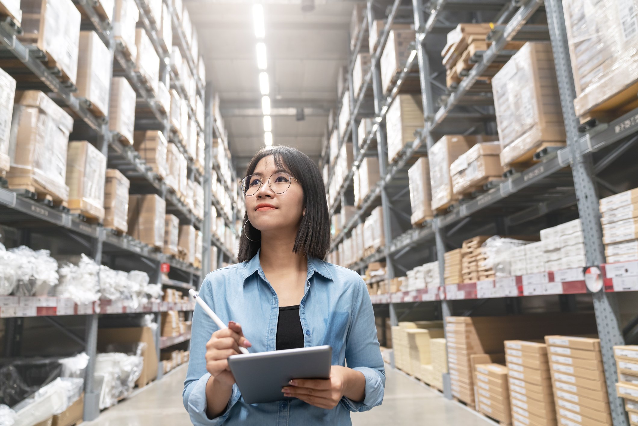 Young woman walking in an aisle of a warehouse as she notes what she sees on her tablet.