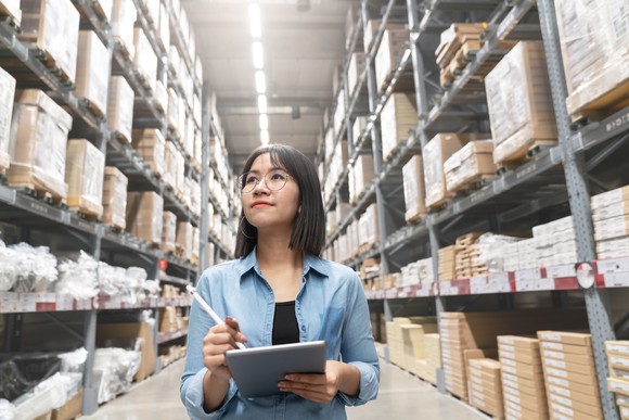 Young woman walking in an aisle of a warehouse as she notes what she sees on her tablet.