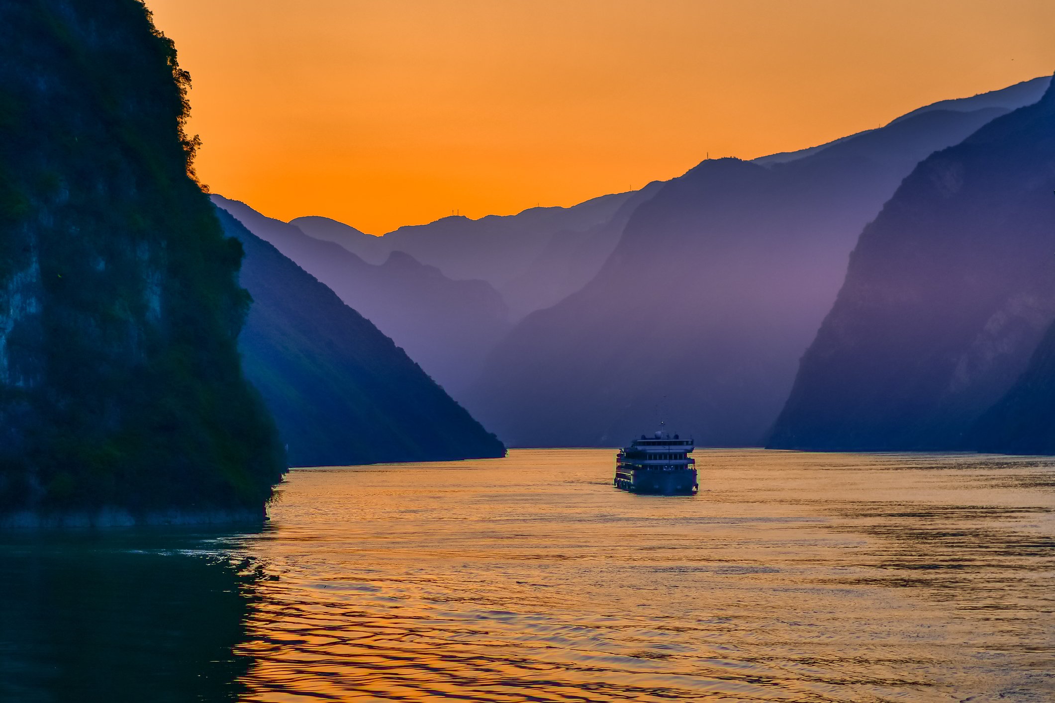 A cruise ship goes into a river surrounded by mountains at sunset. 