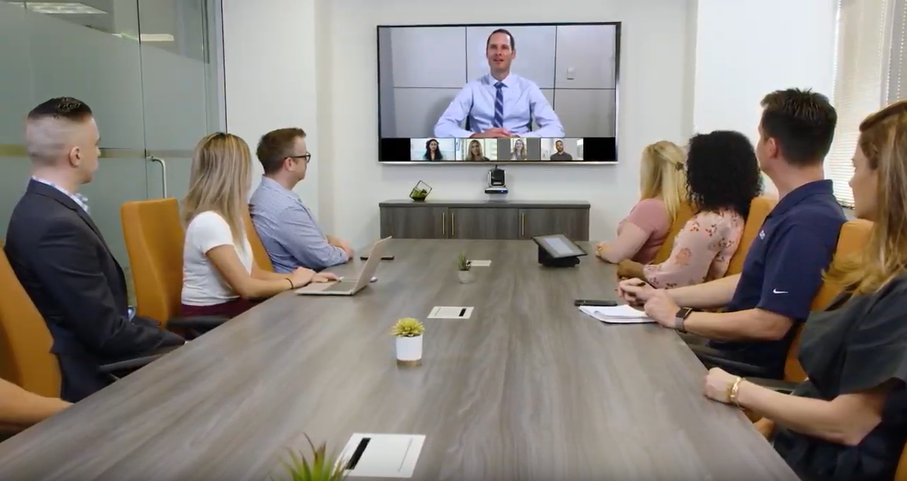 People sitting at a table in a conference room, looking at a person on screen.