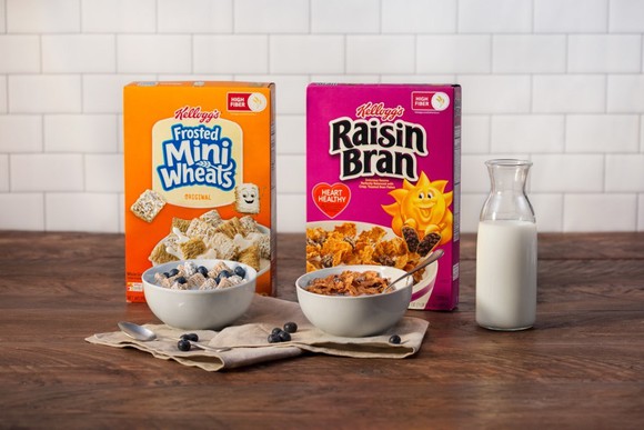 Kellogg cereals Mini Wheats and Raisin Bran sit on a table with bowls and a glass pitcher of milk