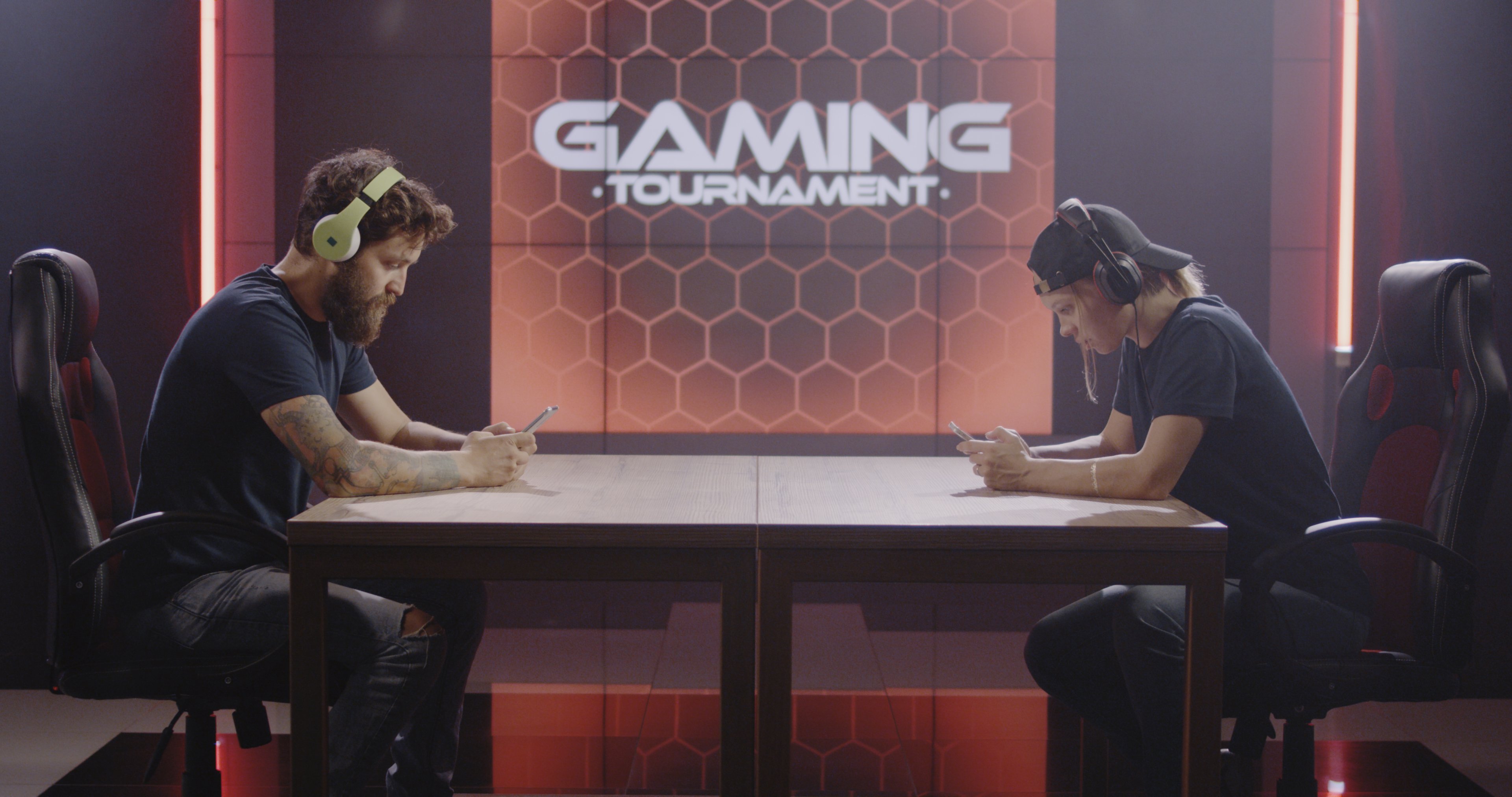 Two men sit across a table holding video game controls with the words Gaming Tournament on a screen behind them.