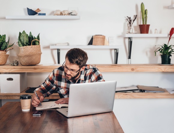 Man taking notes at laptop in what looks like an at-hone arrangement, with plants behind him