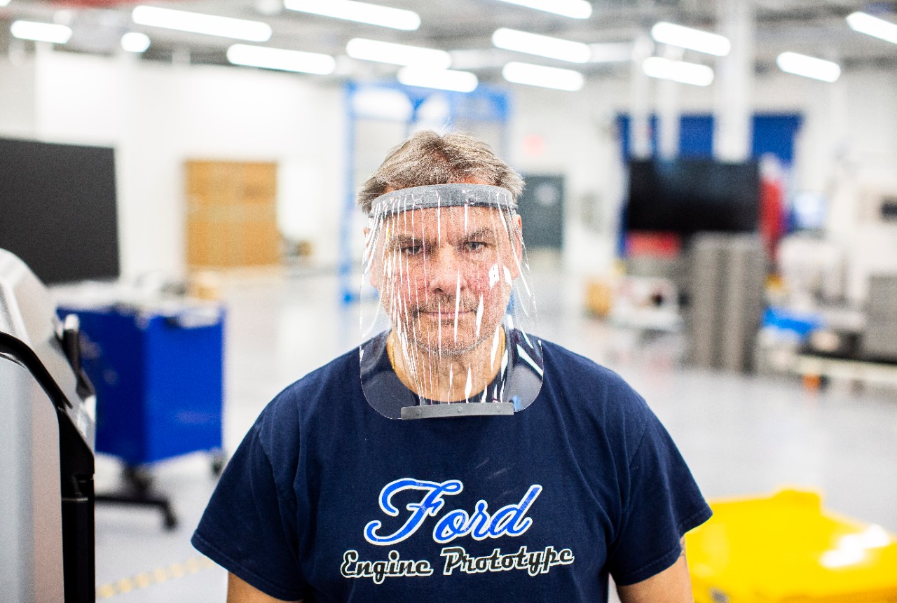 Jacek, wearing a "Ford Engine Prototype" t-shirt and the face shield, is standing on a factory floor.