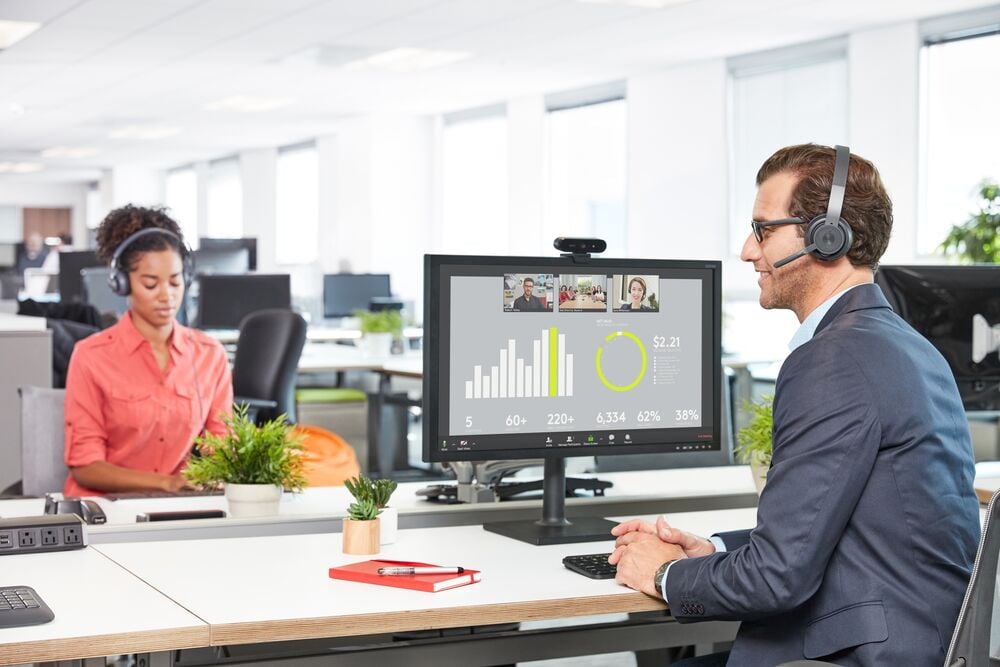 A man sits at an office desk wearing a headset in front of a computer during a video conference.