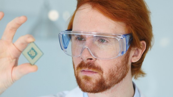A bearded technician wearing goggles holds up a semiconductor chip to take a closer look at it.