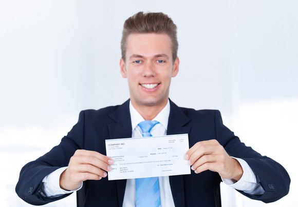 Young man in a suit holds a paycheck and smiles. 